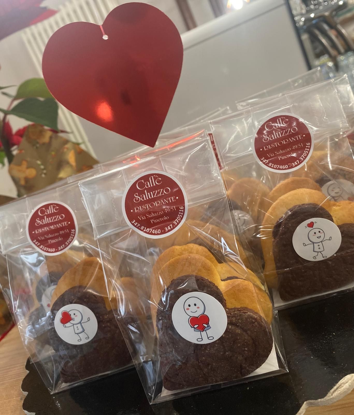Biscotti a forma di cuore in sacchetti trasparenti, decorati con un adesivo a forma di cuore e un'etichetta a forma di cuore rosso appesa sopra.