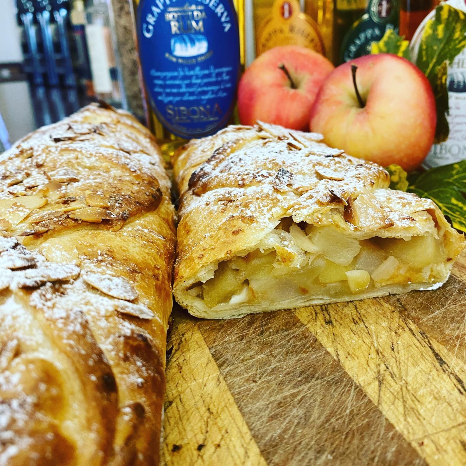 Strudel di mele con mandorle a lamelle e zucchero a velo, con mele e bottiglie sullo sfondo.