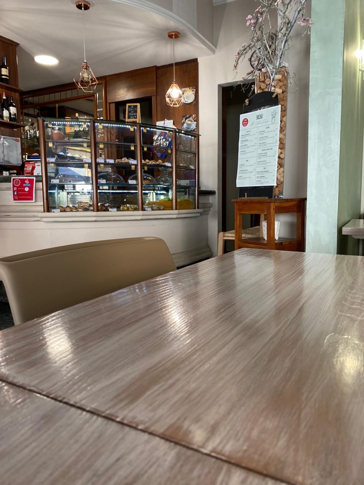 Vista interna di un bar con tavolo in legno, menù, esposizione di dolci e luci decorative.
