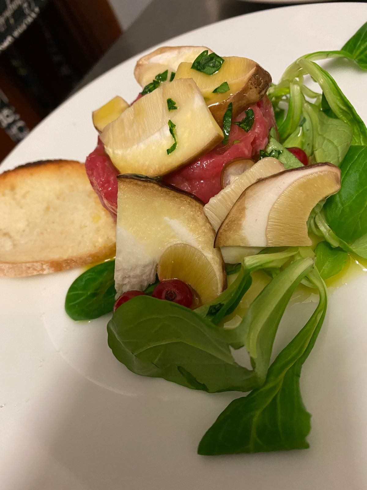 Tartare di manzo con funghi affettati, verdure e pane tostato su un piatto bianco.