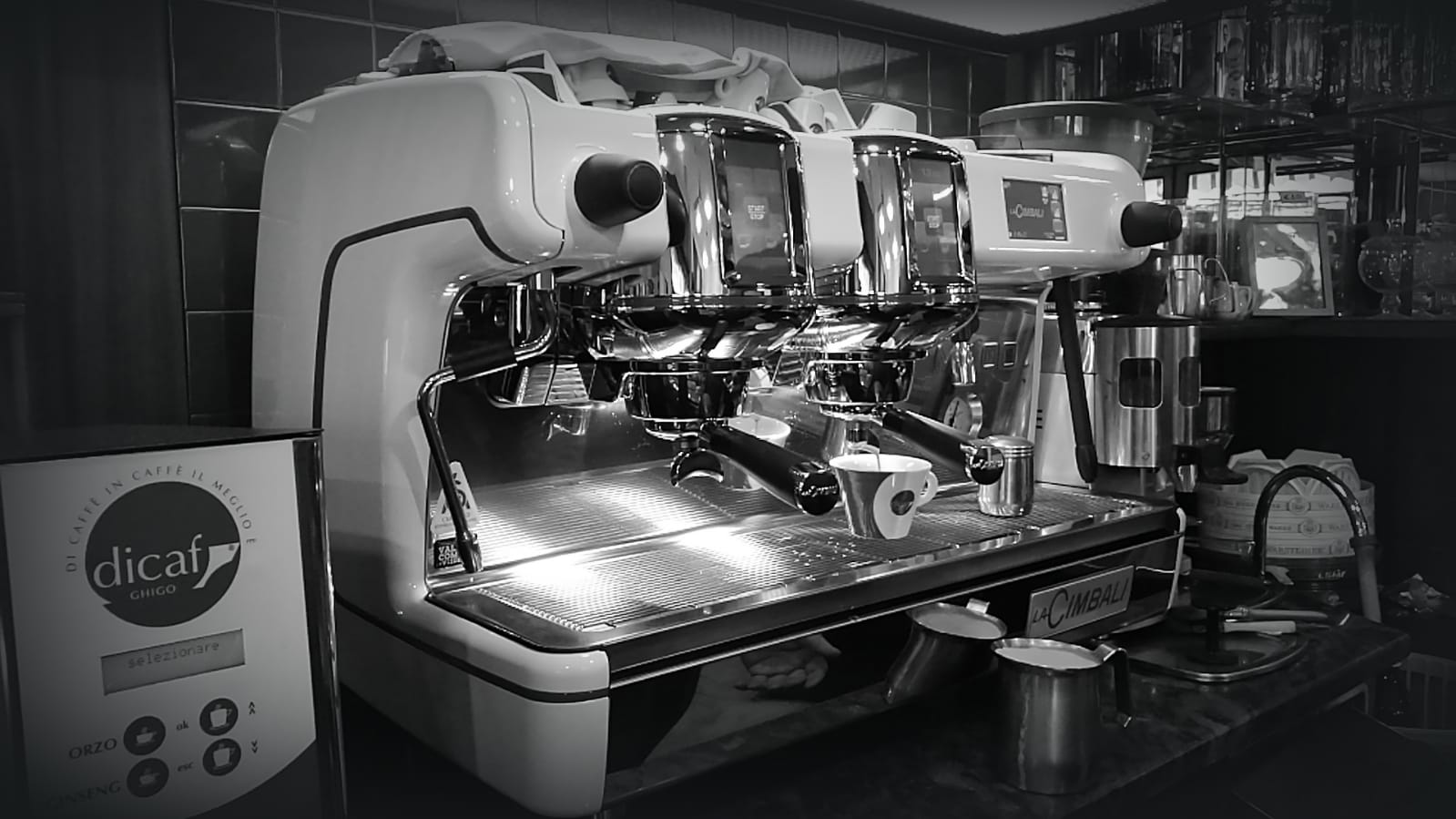 Ripresa in bianco e nero di una macchina per caffè espresso commerciale su un piano di lavoro con comandi digitali e dettagli cromati.