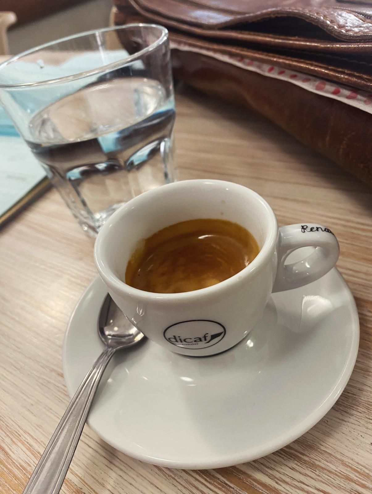 Espresso in una tazza bianca su un piattino, con un bicchiere d'acqua, su un tavolo di legno chiaro.