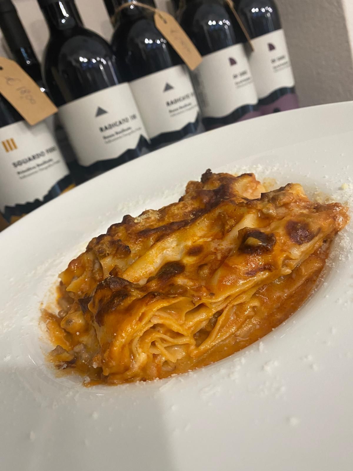 Lasagna su un piatto bianco, con bottiglie di vino sullo sfondo.