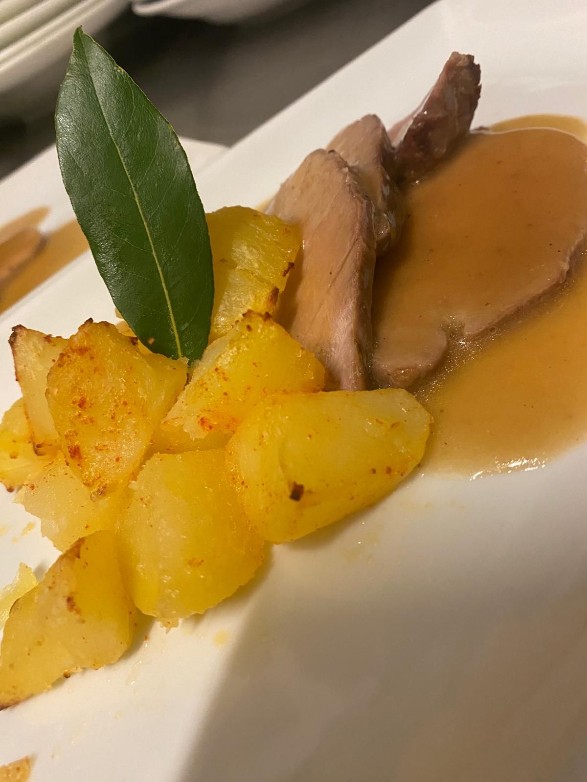 Maiale arrosto con patate e sugo, guarnito con una foglia di alloro, su un piatto bianco.