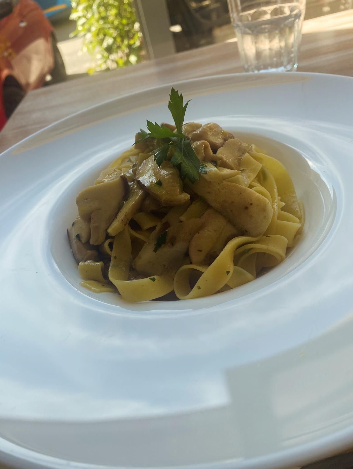 Piatto di pasta con funghi e prezzemolo su un piatto bianco, vicino a un bicchiere d'acqua.