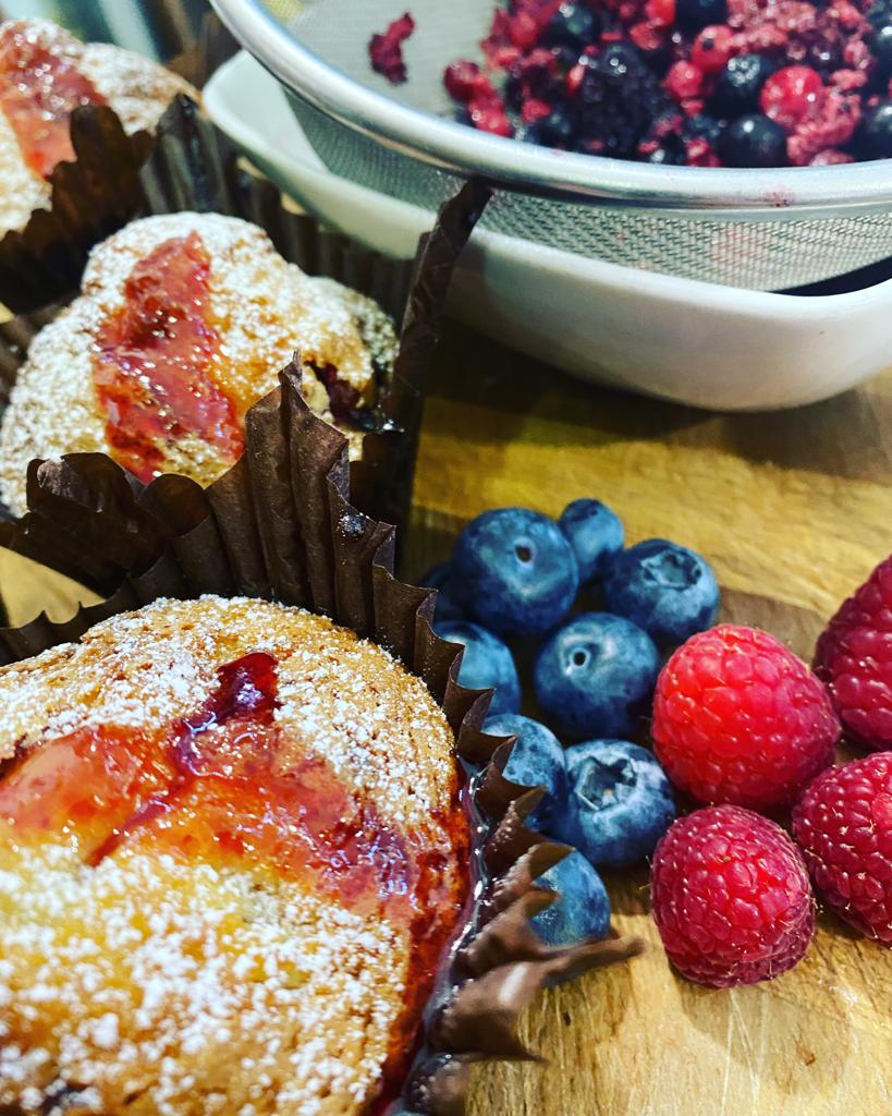 Muffin con marmellata e zucchero a velo, accanto a mirtilli, lamponi e una ciotola di frutti di bosco misti.