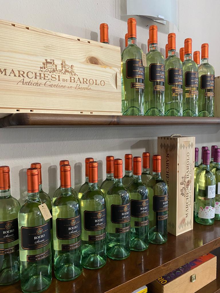 Bottiglie di vino sugli scaffali, alcune con tappi arancioni. Sullo sfondo una scatola di legno e una parete chiara.