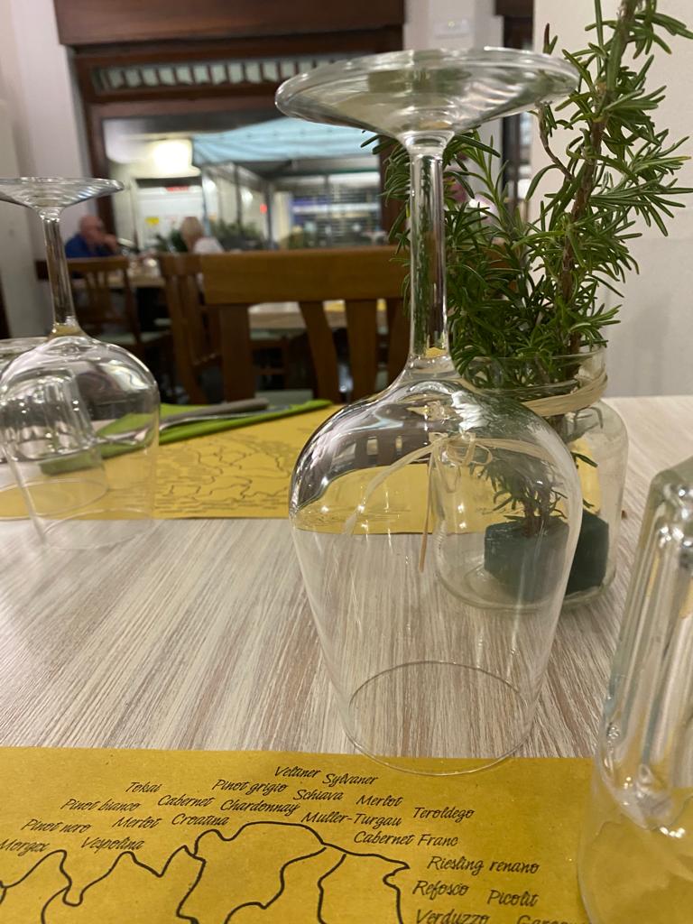 Bicchieri da vino su un tavolo apparecchiato per la cena, rametti di rosmarino in un vaso e l'interno di un ristorante sullo sfondo.