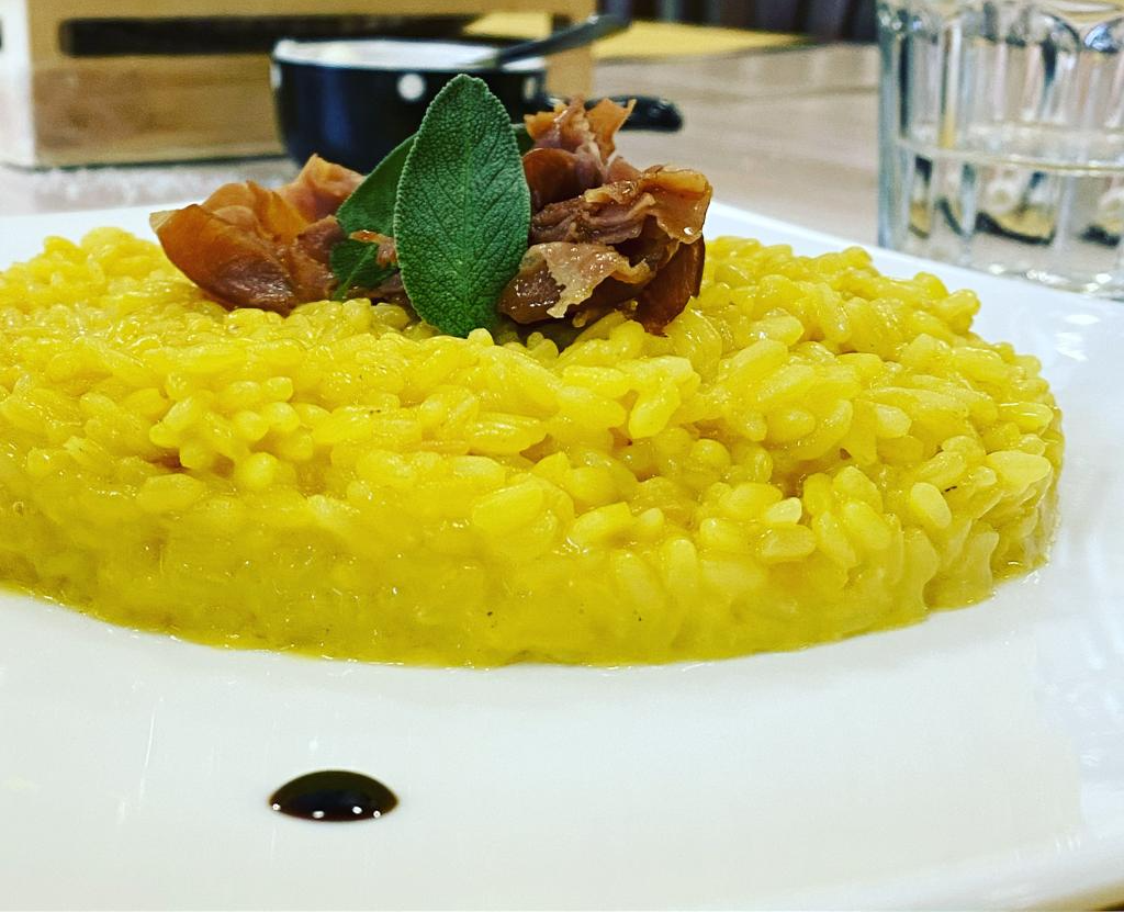 Risotto giallo con prosciutto, una foglia di salvia e glassa di aceto balsamico su un piatto bianco.