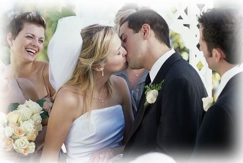 Bride and groom kissing at a wedding, with a smiling bridesmaid holding a bouquet nearby