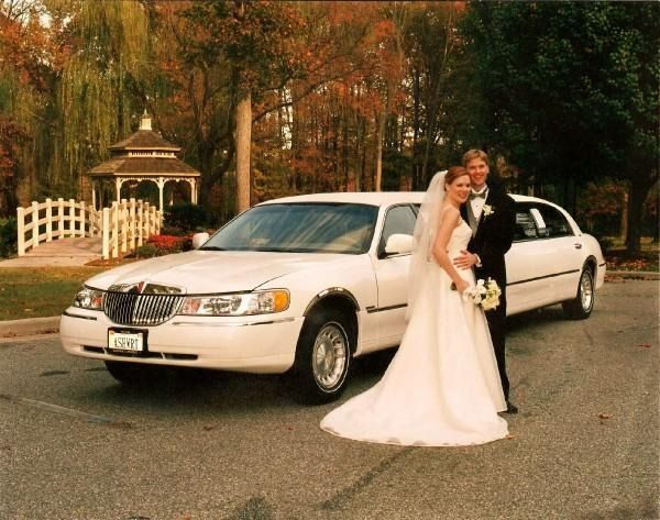 Bride and groom beside a white limousine on a tree-lined street, with autumn leaves and a gazebo in the background