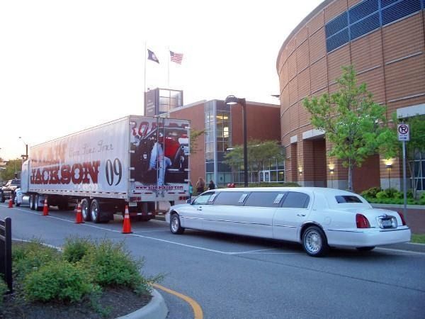 White stretch limousine parked beside a large building and tour truck with orange cones on a city street.