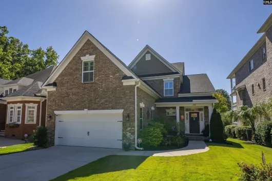 Suburban brick house with white garage, front porch, and manicured lawn on a sunny day