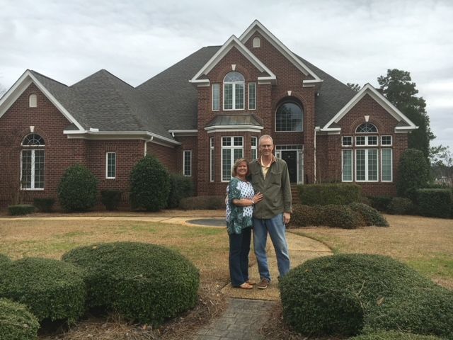 A couple standing in front of a large brick house with manicured bushes and a lawn.