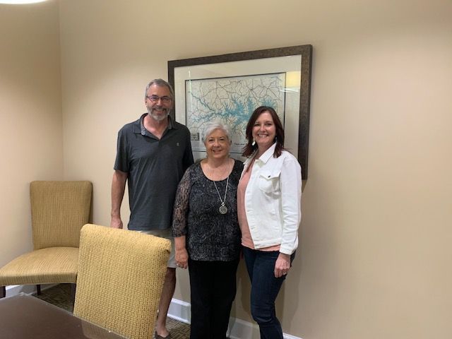Three people standing indoors beside framed artwork and beige chairs