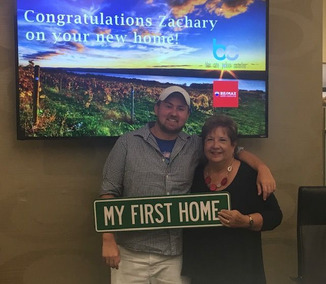 Two people holding a “MY FIRST HOME” sign in front of a congratulatory homeownership display