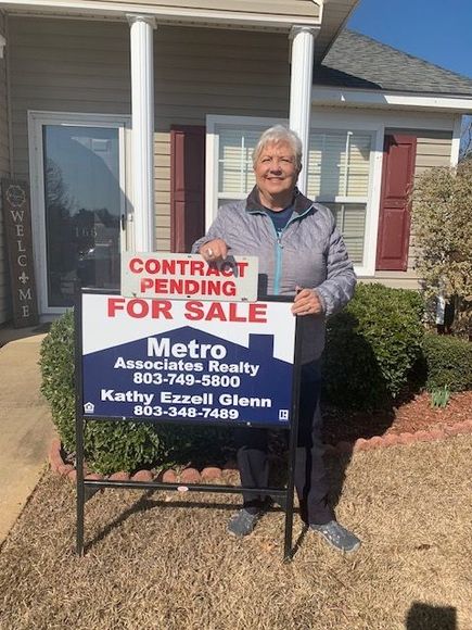 Woman holding a real estate sign that reads “Contract Pending / For Sale” in front of a house