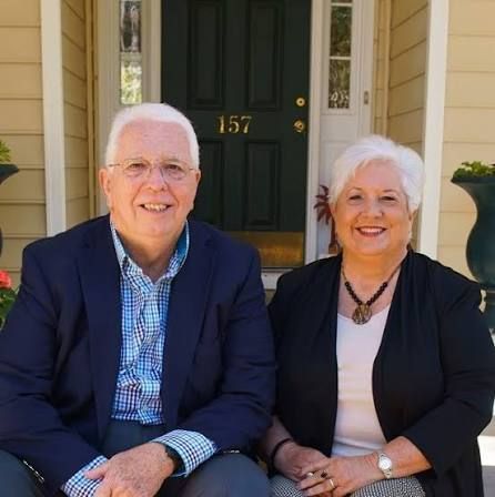 Two smiling people seated on a porch in front of a green door with the number 157
