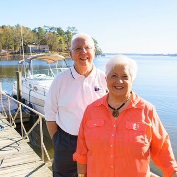 Two smiling people standing by a dock with a boat on a sunny lakefront