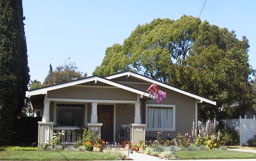 Tan bungalow with white trim, porch, and flowers in the yard.