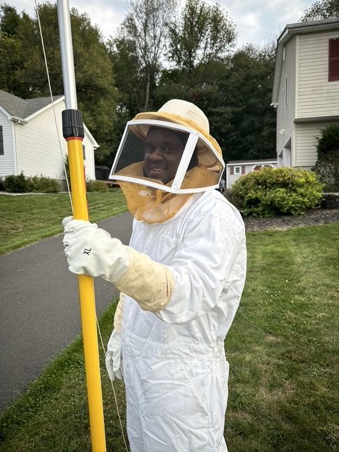 Person in white beekeeping suit holding a yellow pole in a grassy suburban yard