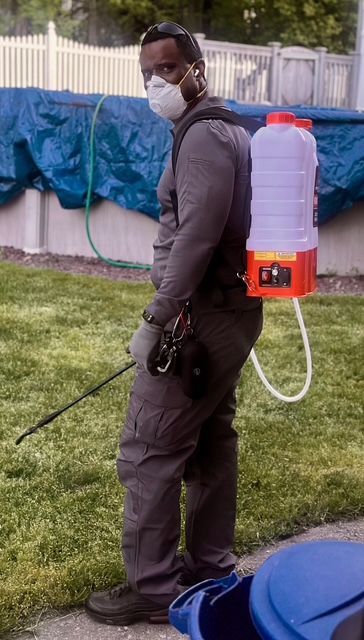 Worker in a mask and brown coveralls spraying yard with a backpack tank sprayer