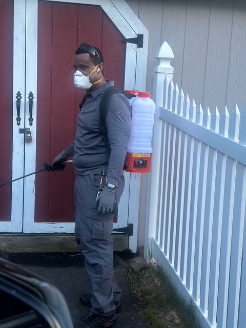 Person in gray workwear and mask standing beside a red shed and white fence, holding a sprayer.