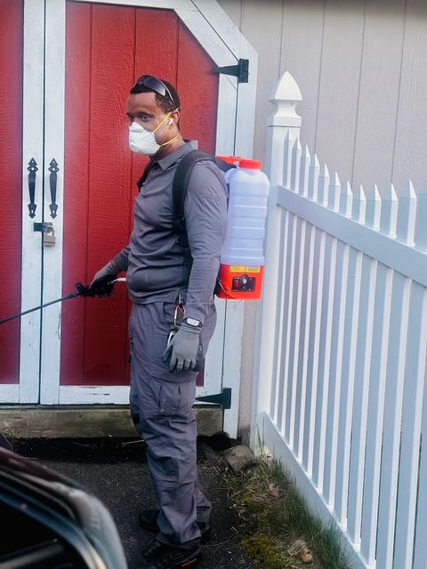 Person in a mask and gloves, carrying a sprayer beside a red shed and white fence.