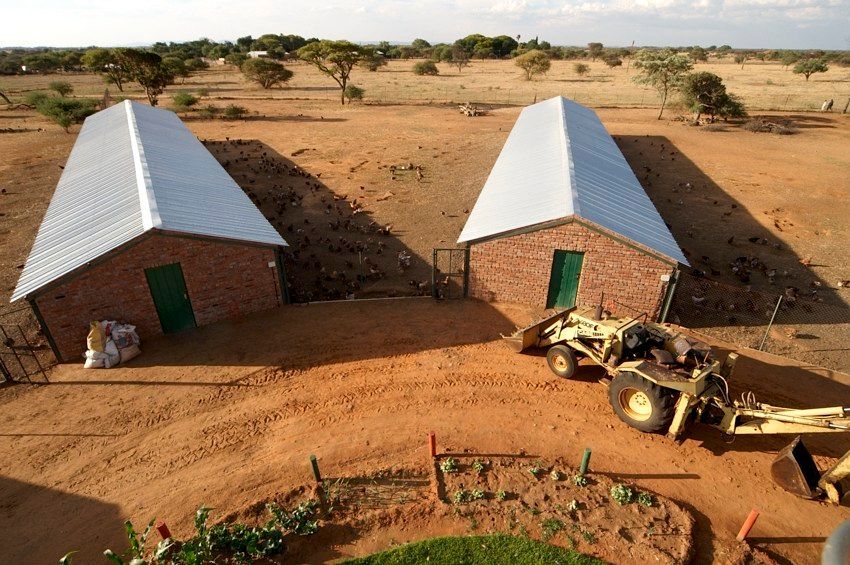 Two small stone huts with metal roofs in a dry rural landscape, with a tractor nearby.