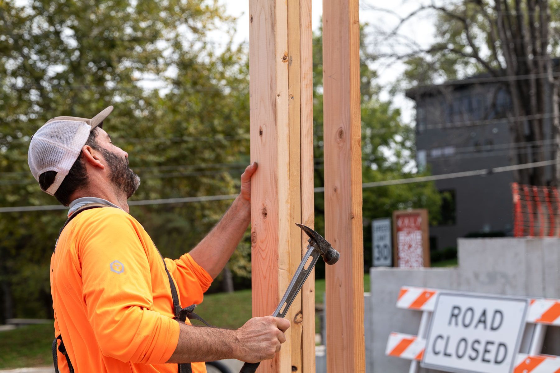 Construction worker in orange shirt and cap hammering wood near a road closed sign.
