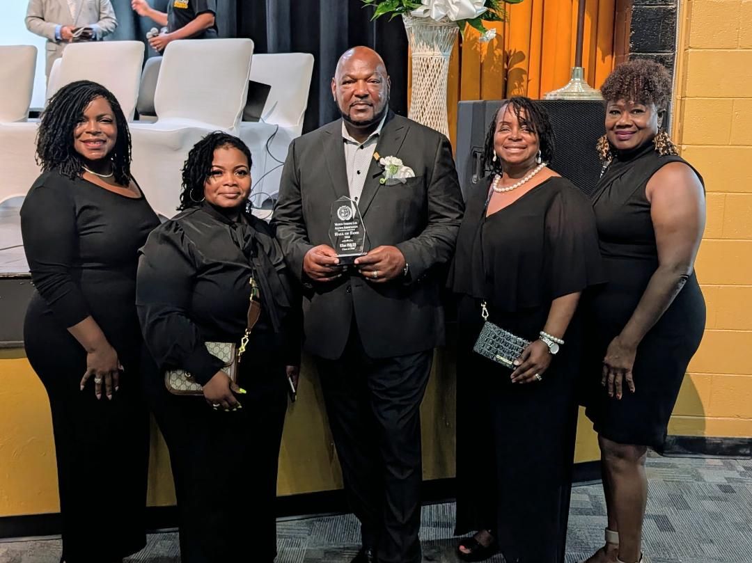 Five people in black outfits posing together indoors at a formal event