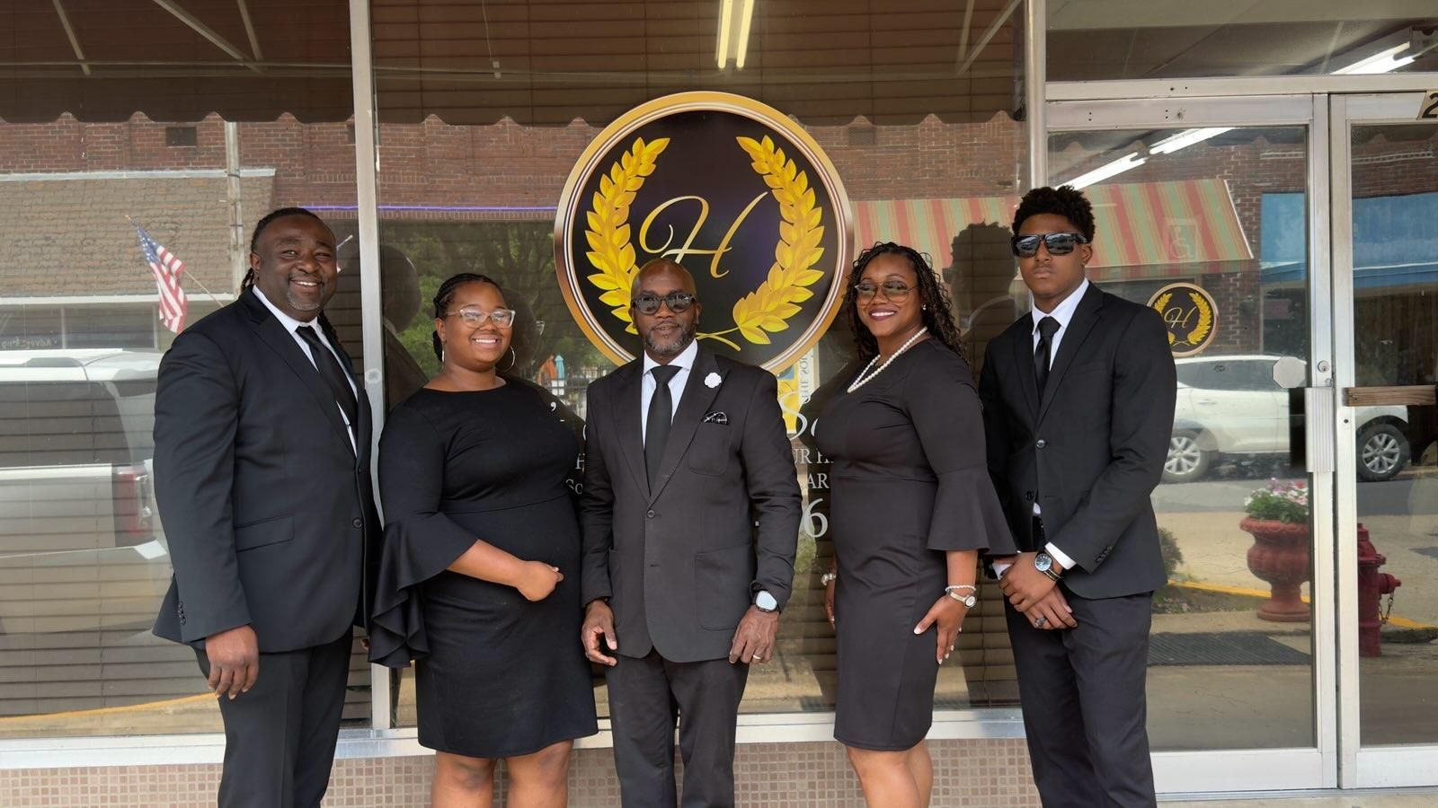 Five people in black formal wear posing in front of a glass entrance with a circular gold logo.