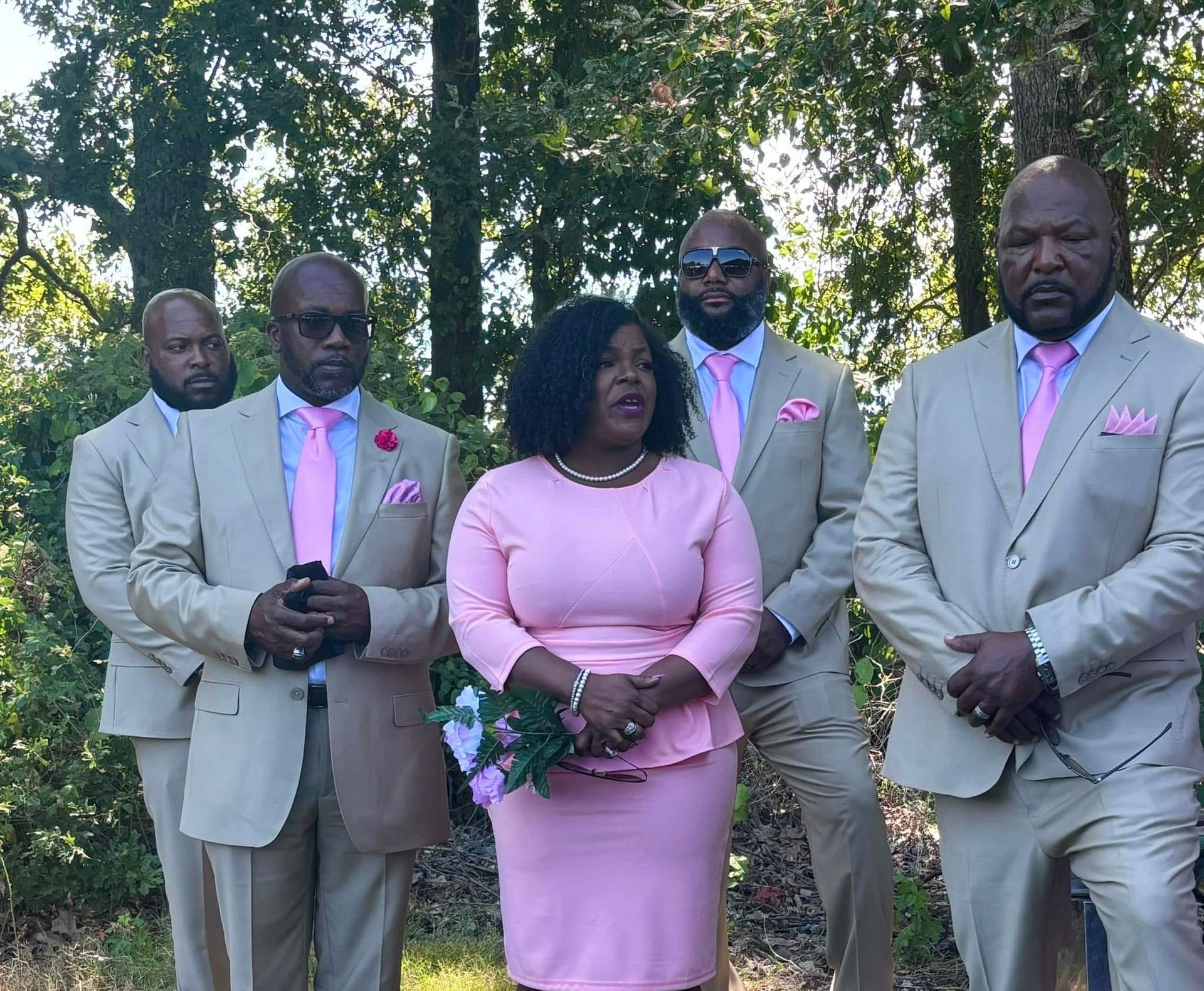 Five formally dressed adults posing outdoors among trees, with pink accents and one woman in a pink dress.