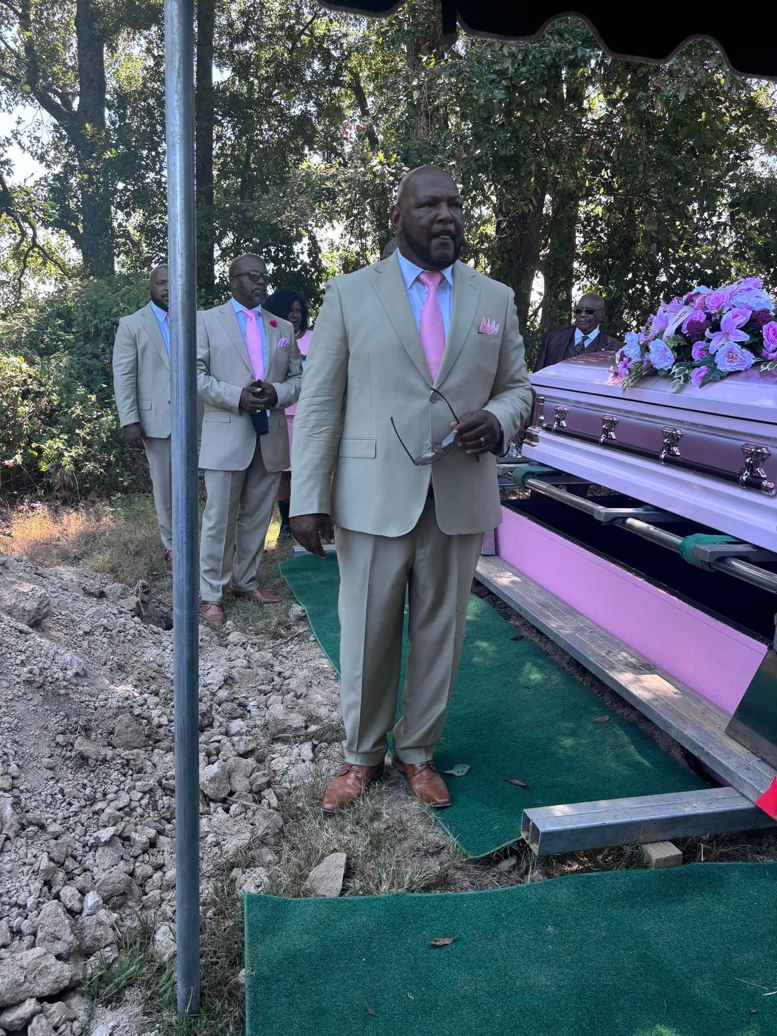 Man in tan suit beside a flower-adorned casket at an outdoor graveside service