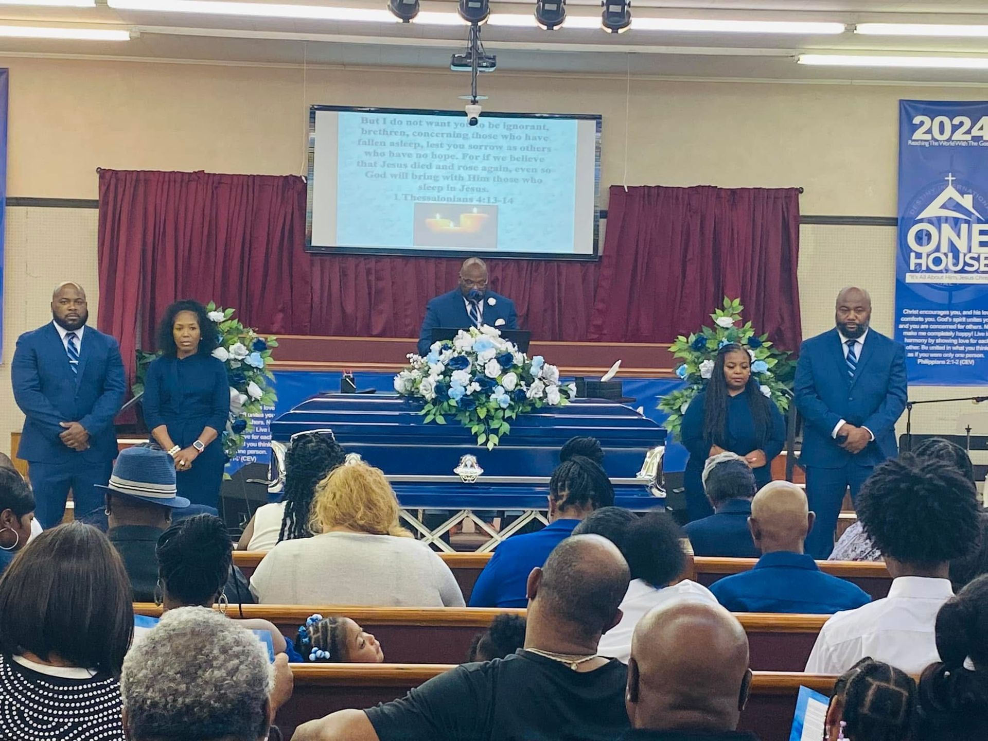 Church service with speaker at podium, blue stage décor, floral arrangement, and seated audience.