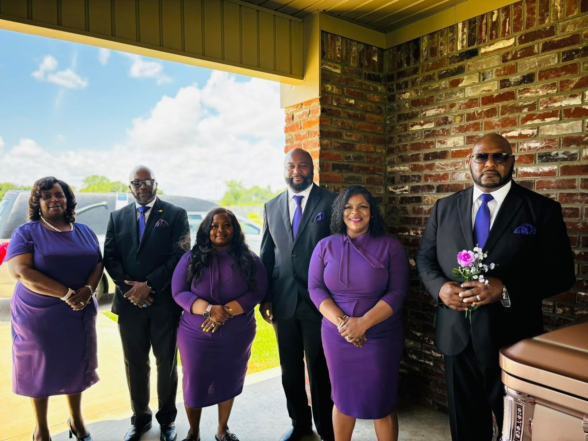 Six formally dressed people in purple and black stand under a covered pavilion outdoors.