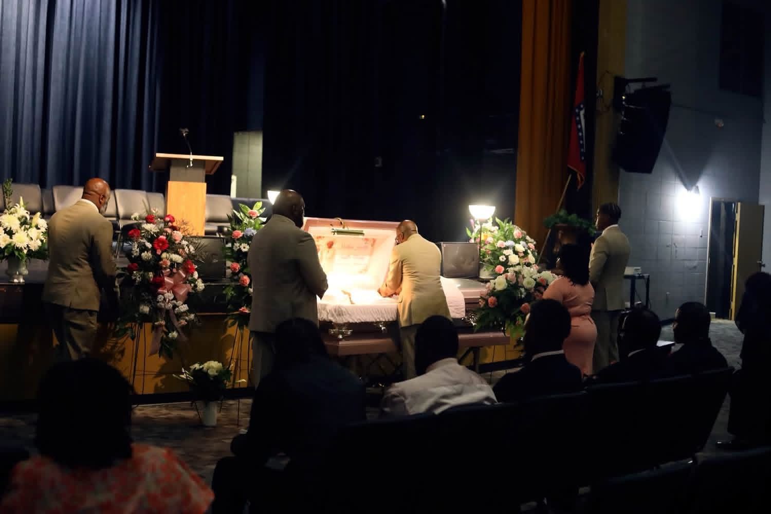 People gathered around a flower-decorated casket on a stage during a funeral service.