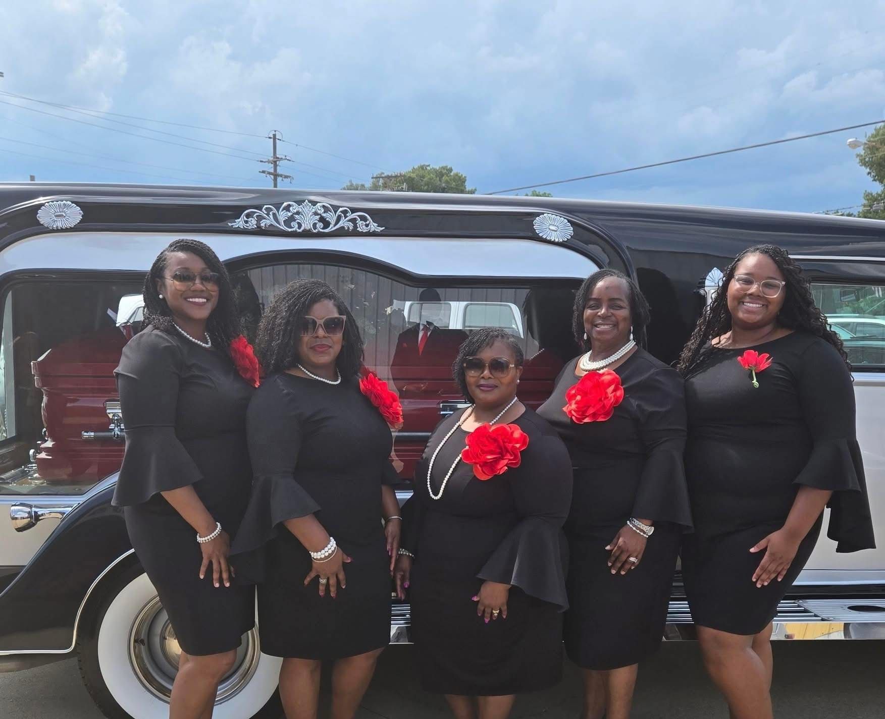 Five women in black dresses with red flowers pose in front of a black car outdoors.