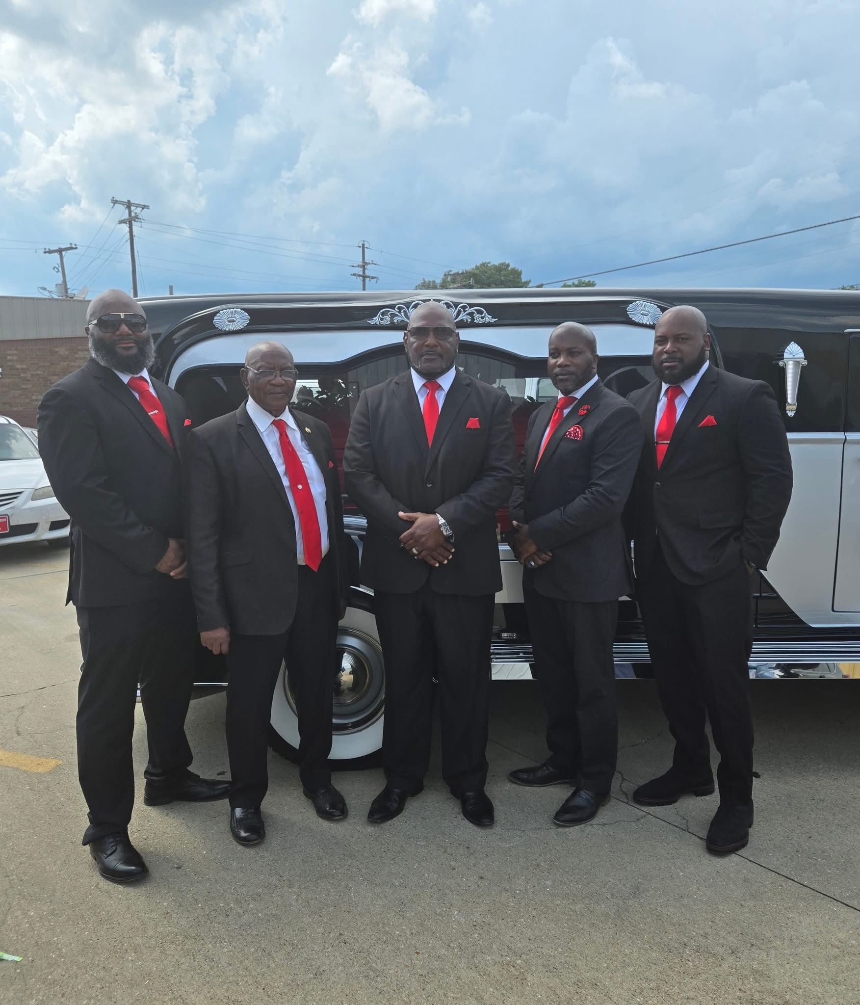 Five men in black suits with red ties pose beside a black van outdoors under cloudy skies