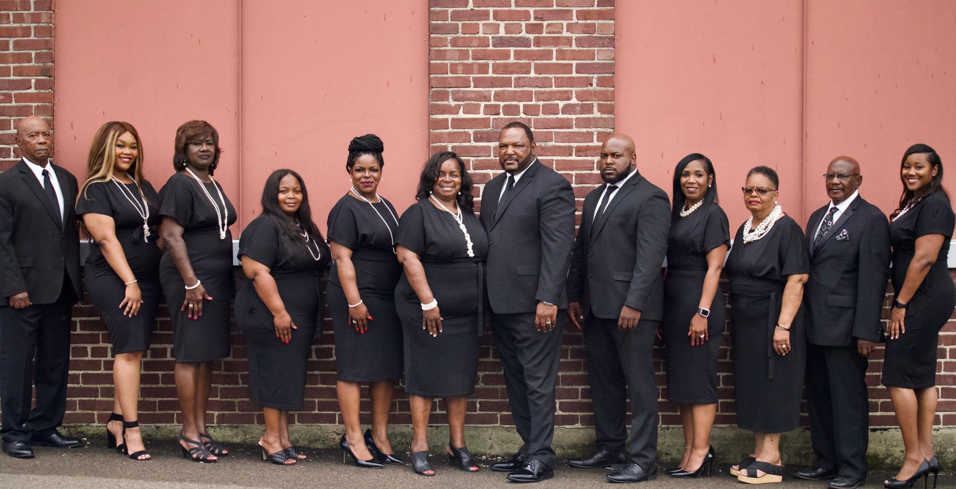 Group of people in black formal attire posing outdoors in front of a brick wall.