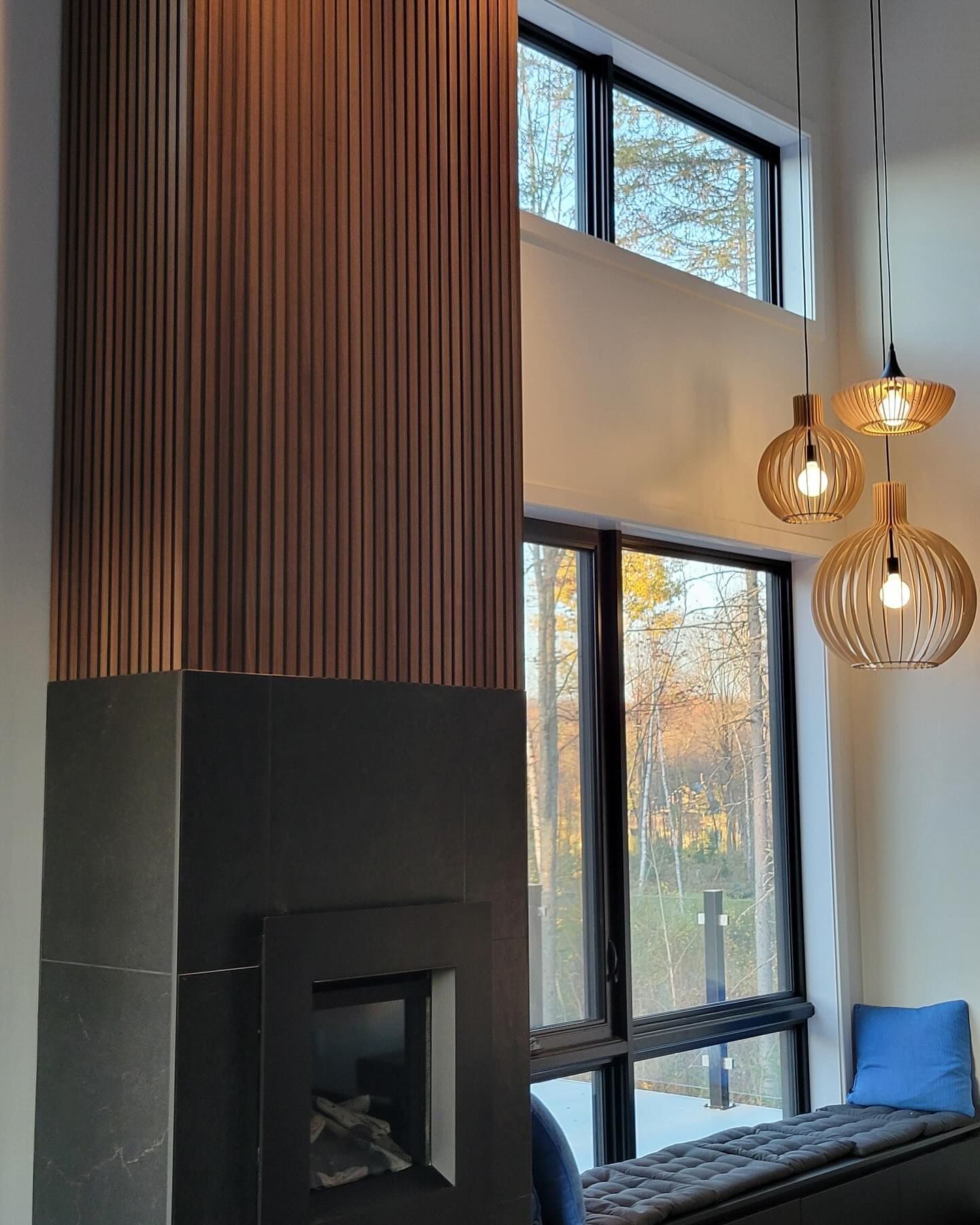 Cheminée moderne avec une base carrelée foncée, entourée de boiseries verticales, à côté d'une grande fenêtre et de suspensions décoratives.