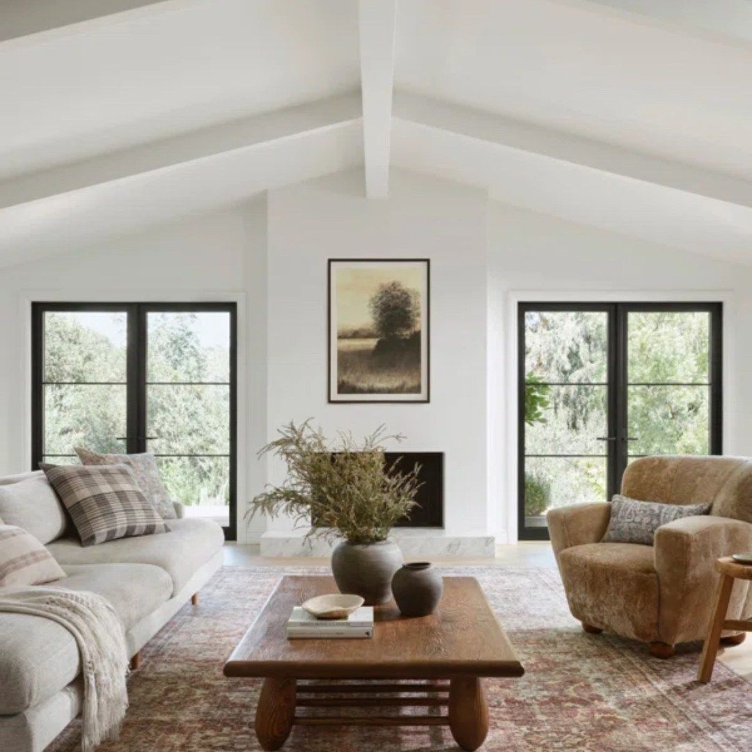 Salon confortable avec des murs blancs, des fenêtres à cadre noir et une table basse rustique.