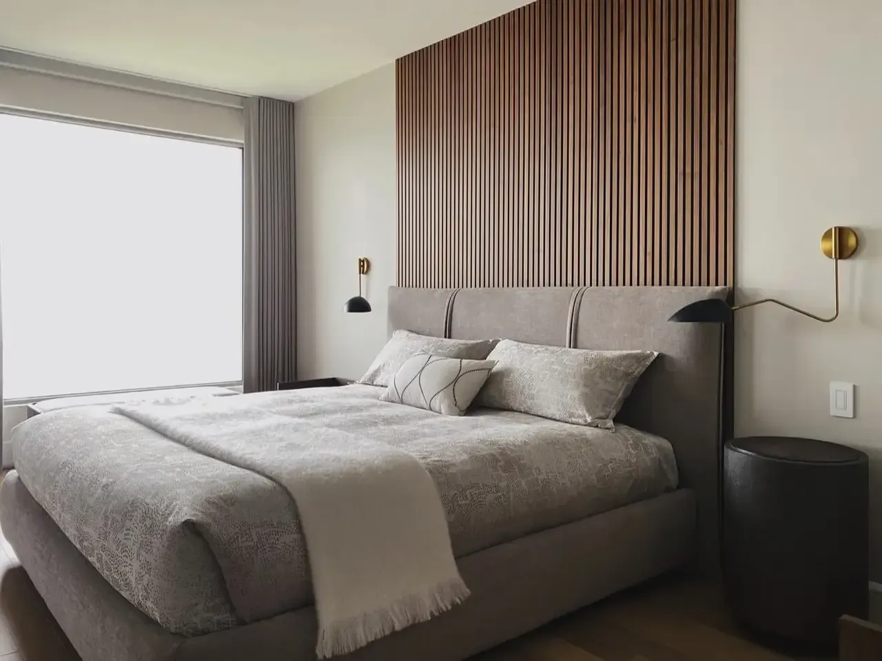Chambre avec un lit contre un mur en panneaux de bois, avec grande fenêtre et deux appliques sur les murs.