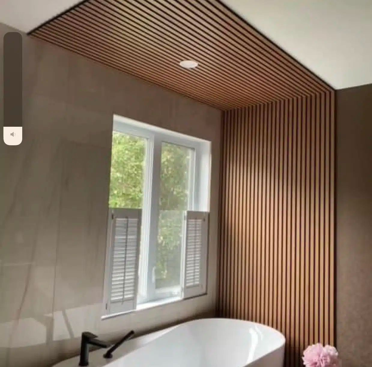Salle de bain avec plafond et mur en lattes de bois, une baignoire blanche et une fenêtre avec volets.