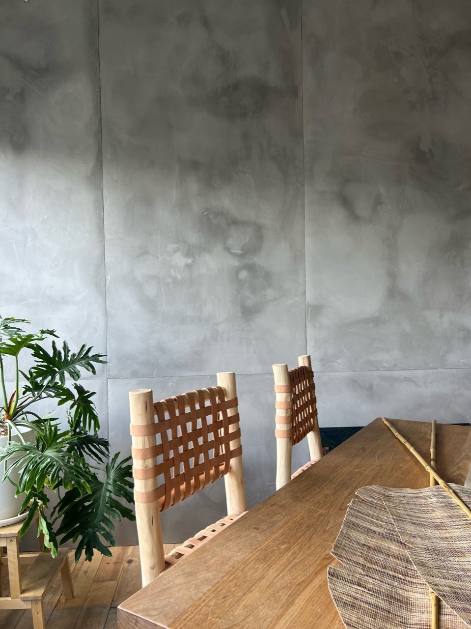 Deux chaises à dossier tressé sont disposées devant une table en bois, contre un mur en béton. Une plante trône dans le coin inférieur gauche.