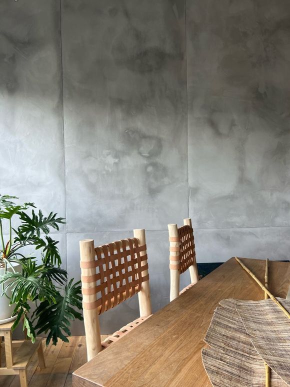 Deux chaises à dossier tressé sont disposées devant une table en bois, contre un mur en béton. Une plante trône dans le coin inférieur gauche.
