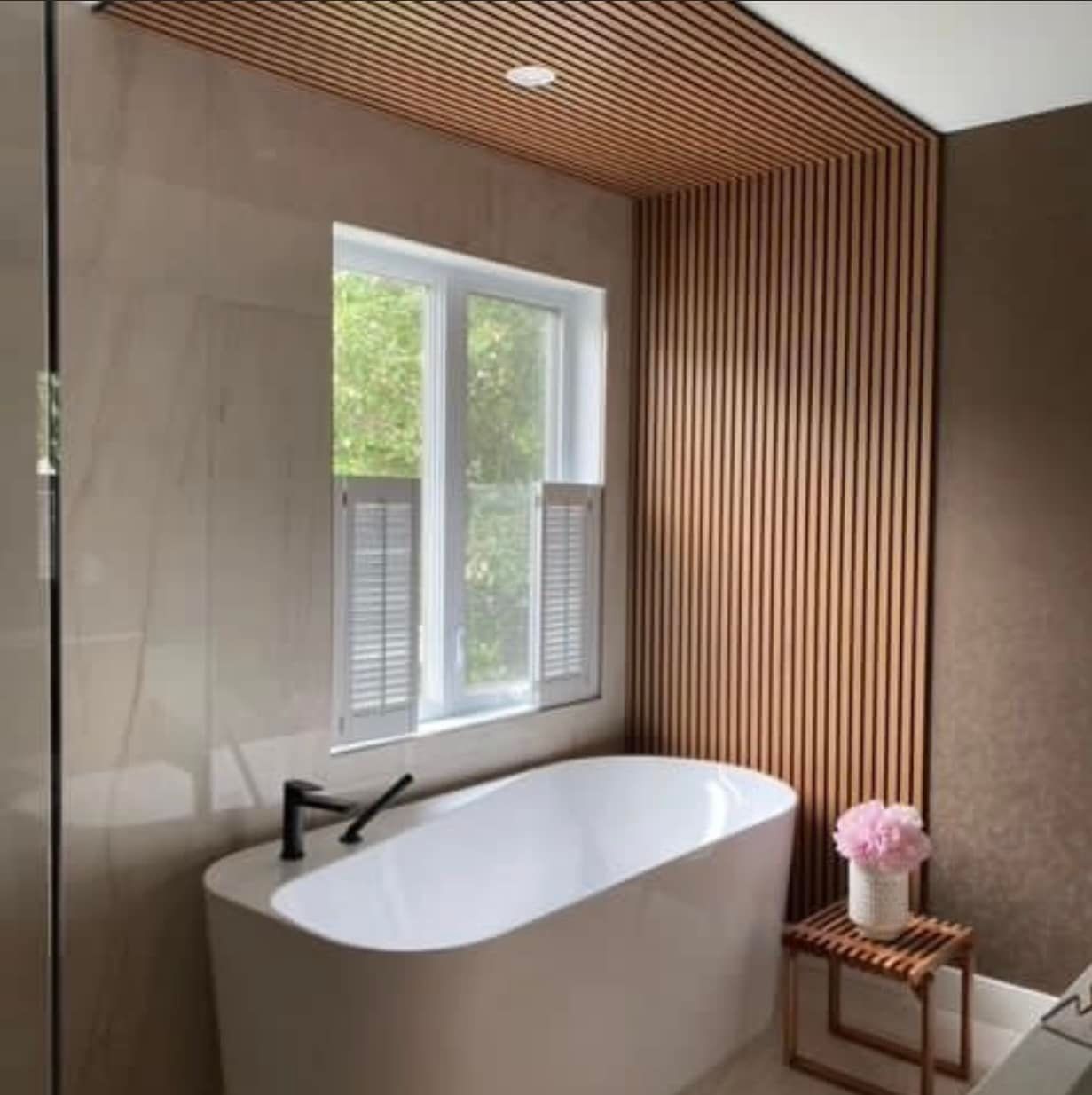 Salle de bain avec baignoire blanche, mur en lattes de bois et fenêtre avec volets.