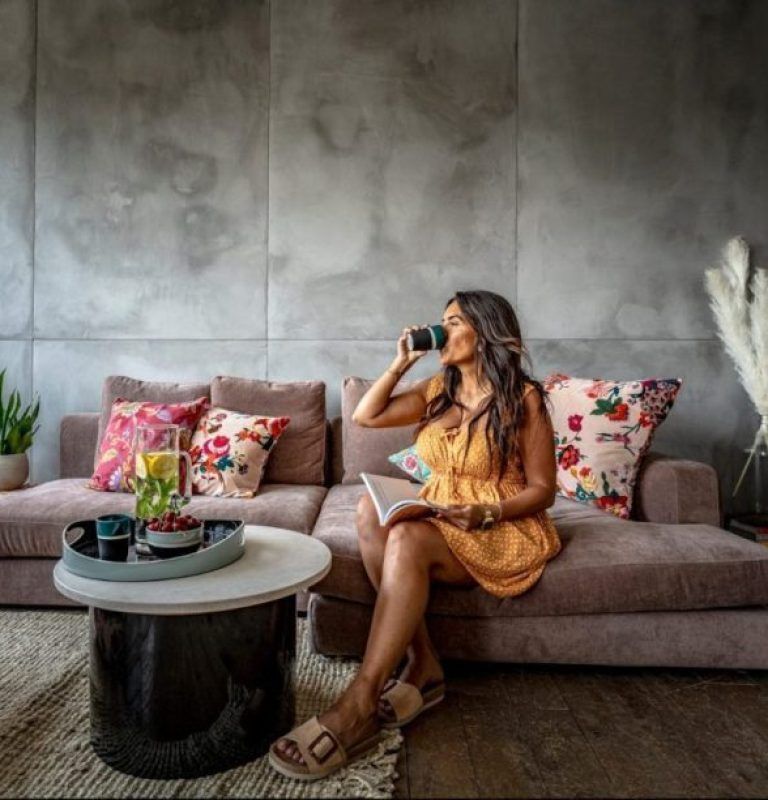Une femme en robe jaune boit du café, assise sur un canapé mauve, un livre à la main. Décor de salon.