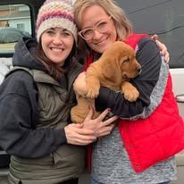 Two women smiling and holding a fluffy brown puppy outdoors by a vehicle.