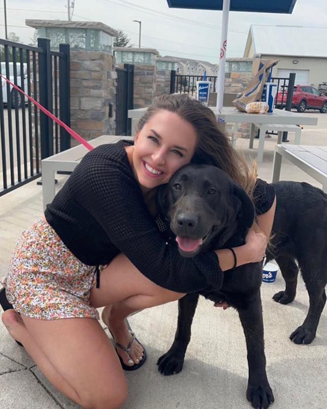 Smiling woman kneels and hugs a black dog on a patio, with another black dog nearby.